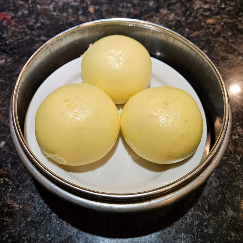 Three golden-yellow steamed buns, likely custard buns, served in a metal dim sum steamer basket.