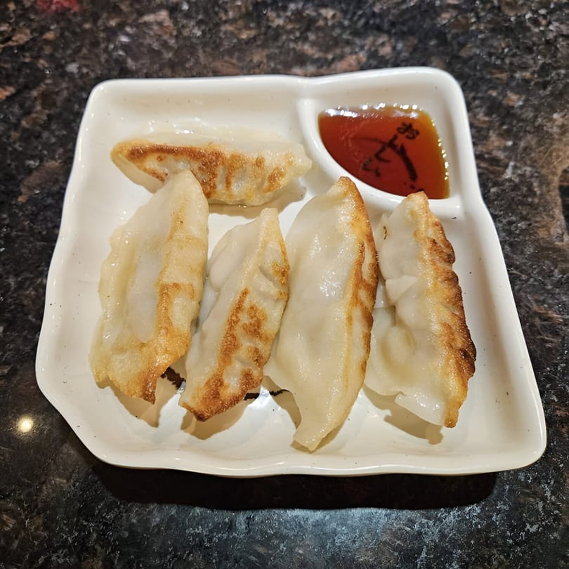 A white rectangular plate of pan-fried dumplings with a side of dipping sauce.