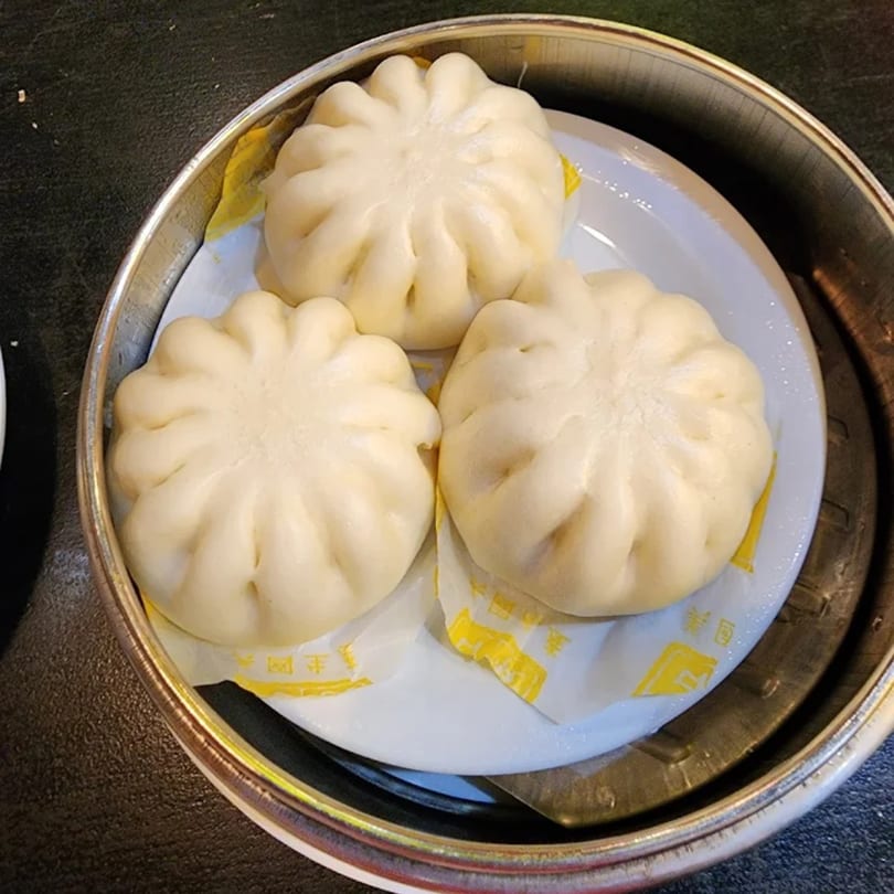Three fluffy steamed buns, likely char siu bao, served in a metal dim sum steamer basket.